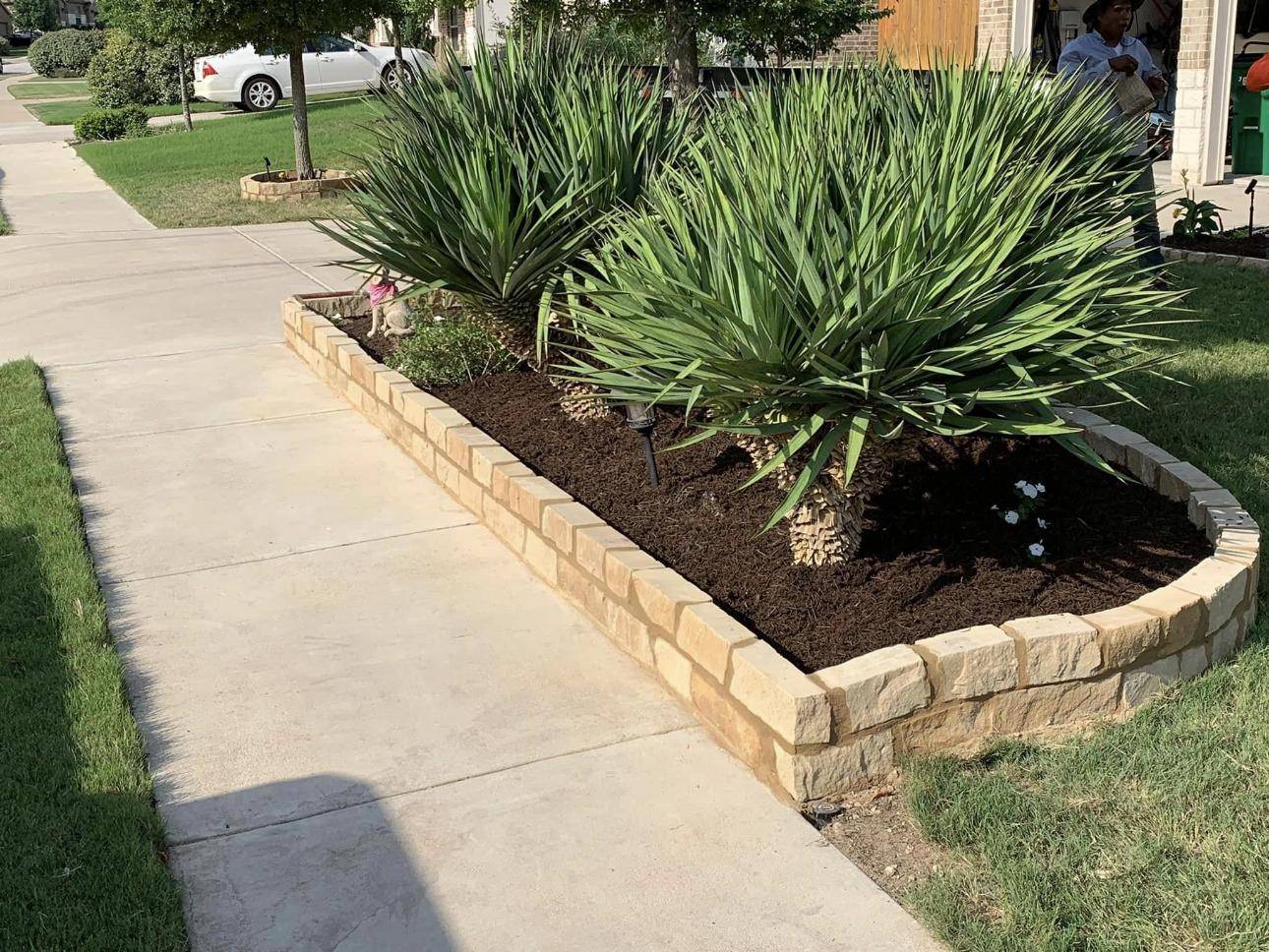 Stone-edged front flower bed with yucca and fresh mulch along a sidewalk