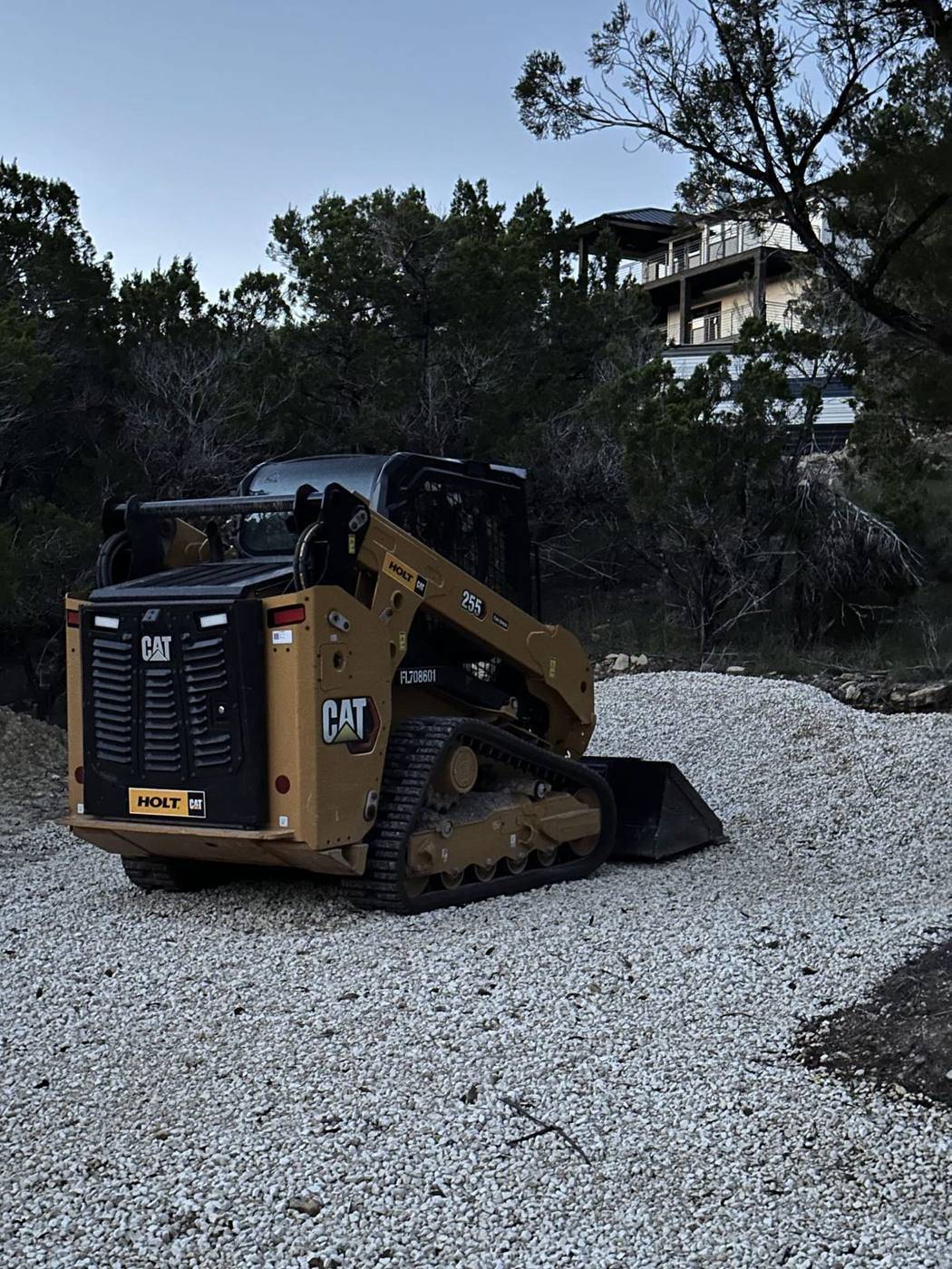 CAT compact track loader spreading crushed limestone gravel on a sloped Hill Country lot below a lake-view home